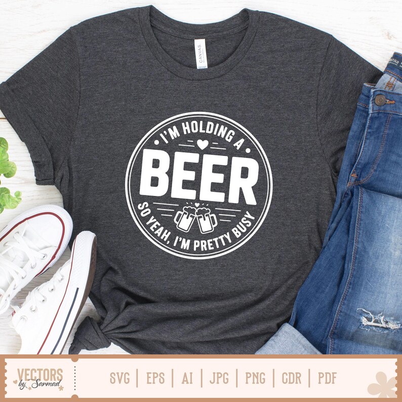 May include: Dark gray t-shirt with a white circular graphic that reads "I'M HOLDING A BEER SO YEAH, I'M PRETTY BUSY." The design includes two beer mugs. The shirt is paired with blue jeans and white sneakers.