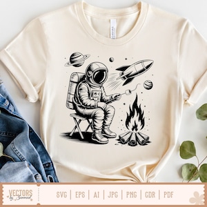 May include: Cream-colored t-shirt featuring a black and white illustration of an astronaut roasting a marshmallow over a campfire. A rocket ship, planets, and stars are also depicted. The shirt has a crew neck and short sleeves.