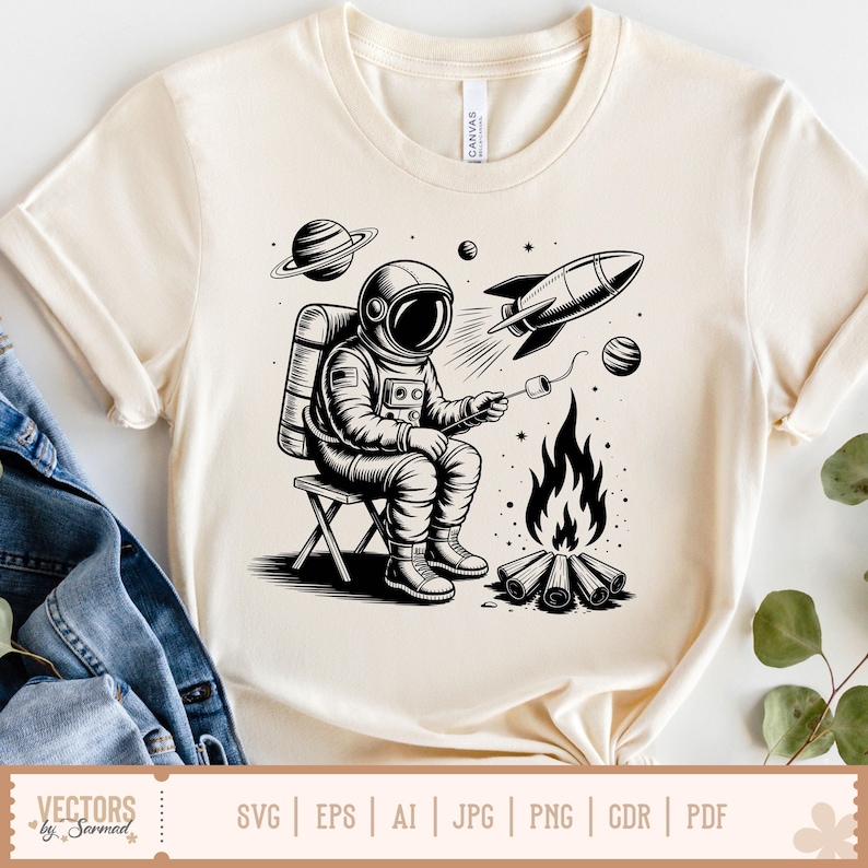May include: Cream-colored t-shirt featuring a black and white illustration of an astronaut roasting a marshmallow over a campfire. A rocket ship, planets, and stars are also depicted. The shirt has a crew neck and short sleeves.