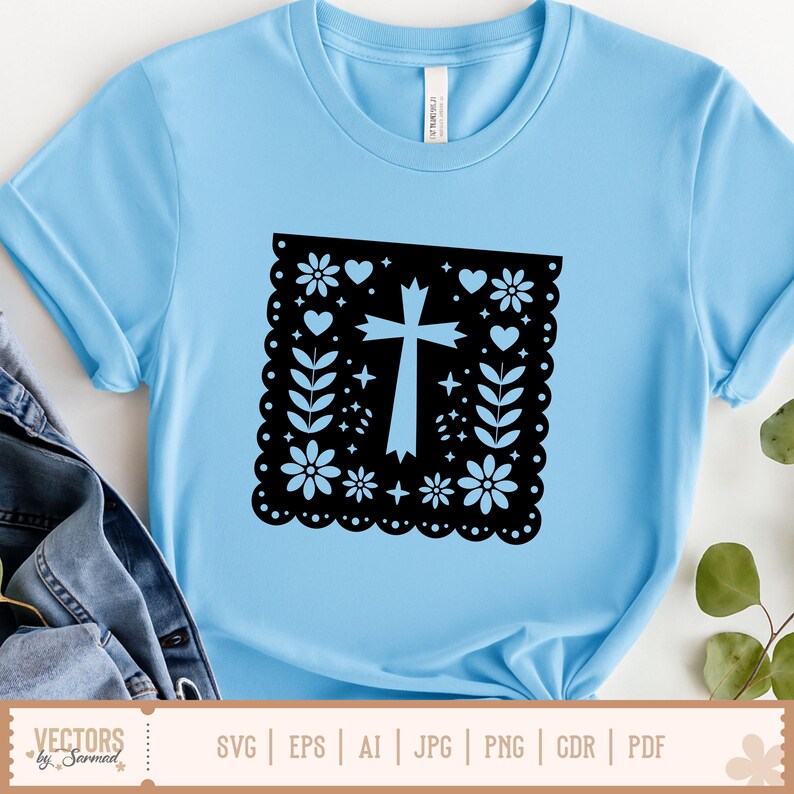 May include: Pale blue t-shirt with a black square design featuring a cross, flowers, hearts, and leaves. The shirt has short sleeves and a crew neck. The design is centered on the front. The shirt is made of a soft material.