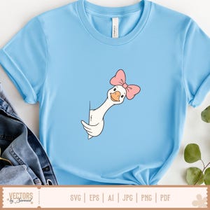 May include: A light blue t-shirt featuring a graphic illustration of a white goose with an orange beak and a pink bow on its head. The goose is depicted peeking out from behind a vertical line, with its neck and head visible in the centre.
