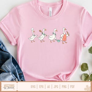 May include: A light pink t-shirt featuring a graphic illustration of four white geese in a row. The geese are depicted with cooking accessories including a chef hat, a wooden spoon, a green neckerchief, a cooking pot, and a coral-coloured apron.