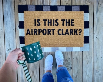 May include: A doormat with the text "IS THIS THE AIRPORT CLARK?" sits on a wooden deck. A green mug with white tree designs is held near the mat. The mat has a tan center and black and white checkered borders.