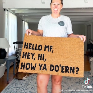 Puede incluir: Una alfombra de coco marrón con el texto "HELLO MF, HEY, HI, HOW YA DO'ERN?" en letras negras.