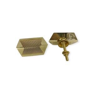 May include: Two gold cabinet knobs with a geometric design. One knob has a textured centre and the other has a screw for installation. The knobs are hexagonal and have a shiny finish. The knobs are likely for furniture.