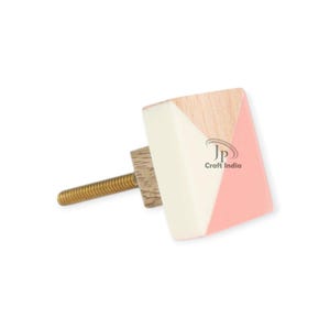 May include: A decorative cabinet knob with a geometric design. The knob features a square shape with sections of light wood, white, and pink. A gold-coloured screw extends from the back. The text "Craft India" is visible.