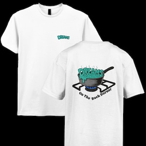 May include: White t-shirt with the word "DREAMS" in teal on the front and back. The back features a graphic of a pan with "DREAMS" in teal, overflowing onto a stove burner, with the text "On The Back Burner".