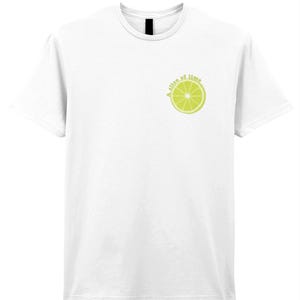 May include: White t-shirt with a crew neck and short sleeves. The shirt features a lime slice graphic with the text "A slice of time" in a light green color on the upper left chest area. The shirt is made of a soft material.