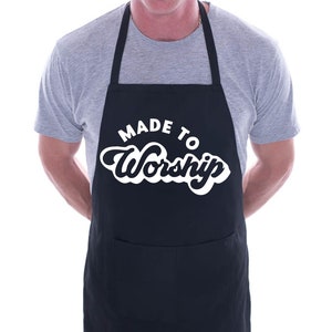 Op de afbeelding: Een zwarte schort met een witte zak en de tekst "Made to Worship" in een wit retro-lettertype.