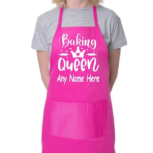 May include: Pink apron with a white graphic that says "Baking Queen" with a crown and rolling pins. The text "Any Name Here" is below the graphic.