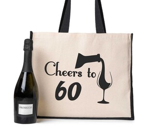 Op de afbeelding: Een crèmekleurige canvas draagtas met zwarte handvatten en randen, naast een fles Prosecco. De tas heeft de tekst "Cheers to 60" en een afbeelding van wijn die wordt geschonken. De fles is donkergroen met een wit etiket.