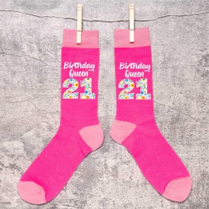 Peut inclure: Deux paires de chaussettes rose vif avec l'inscription "Birthday Queen" et le chiffre "21" dans un motif floral. Les chaussettes sont suspendues à une corde à linge avec des pinces à linge en bois. Les chaussettes ont des talons et des orteils roses.