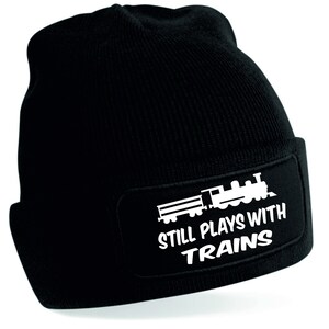 Peut inclure: Bonnet noir en tricot avec un patch blanc représentant un train de dessin animé et le texte "Still Plays With Trains".