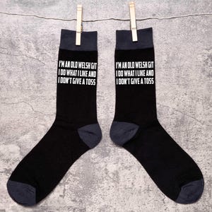 May include: A pair of black socks with a white text design that reads "I'm an old Welsh git I do what I like and I don't give a toss".