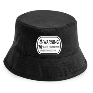 May include: Black bucket hat with a white rectangular patch. The patch features a skull and crossbones symbol and the text "WARNING 70 YEAR OLD GRUMPY GIT MISERABLE, MOODY & FULL OF CRAP."