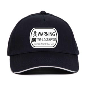 Könnte beinhalten: Schwarze Baseballkappe mit weißem Besatz und einem weißen rechteckigen Aufnäher. Der Aufnäher zeigt einen Totenkopf mit gekreuzten Knochen, das Wort "WARNING" und den Text "80 YEAR OLD GRUMPY GIT". Darunter steht der Satz "MISERABLE, MOODY & FULL OF CRAP".