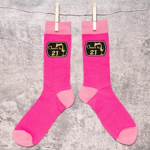 May include: A pair of bright pink socks with light pink heels and cuffs, hanging from wooden clothespins. Each sock features a black and gold label with the text "Cheers to 21" and a wine glass illustration. The socks are displayed against a gray background.