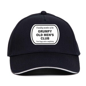Op de afbeelding: Een zwarte baseballpet met witte rand en een witte patch. De patch luidt: "Oprichter van de GRUMPY OLD MEN'S CLUB. Nooit blij tenzij klagen."