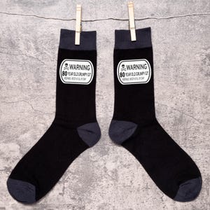 May include: A pair of black socks with dark gray accents, each featuring a white rectangular patch with the text "WARNING 80 YEAR OLD GRUMPY GIT MISERABLE MOODY & FULL OF CRAP" in black. The socks are hanging from a clothesline.