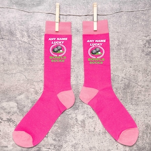 May include: Two pairs of bright pink socks are displayed, each held by a wooden clothespin. The socks have a light pink toe and cuff. The text "ANY NAME LUCKY BOWLS SOCKS" is printed on the cuff, along with a graphic of bowling balls.