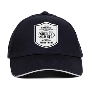 Può includere: Un cappellino da baseball blu navy con finiture bianche e una grafica in bianco e nero. La grafica recita: "I NEVER DREAMED THAT ONE DAY I'D BECOME A GRUMPY OLD GIT BUT HERE I AM PERFECTING IT."