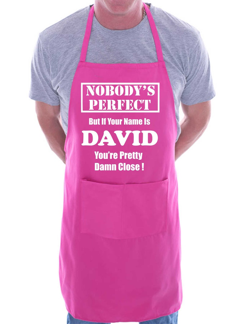 Puede incluir: Delantal rosa con un bolsillo blanco y el texto "Nobody's Perfect But if Your Name is DAVID You're Pretty Damn Close!"