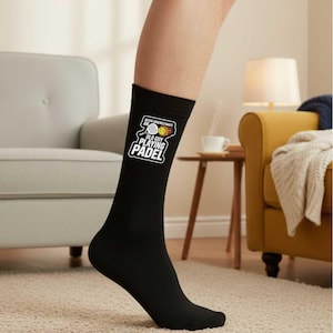 May include: Black crew sock with the text "NEVER UNDERESTIMATE AN OLD GUY PLAYING PADEL" in white. The design includes a padel racket and ball graphic. The sock is worn on a leg.