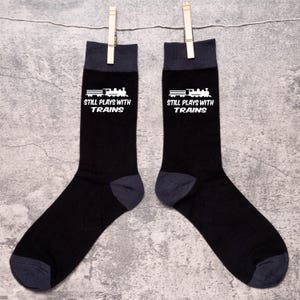 Puede incluir: Un par de calcetines negros con talones, dedos y puños de color gris oscuro. Cada calcetín presenta un gráfico blanco de un tren y el texto "STILL PLAYS WITH TRAINS". Los calcetines cuelgan de un tendedero con pinzas de madera.