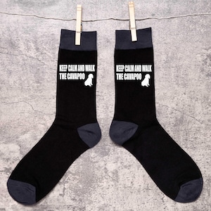 Puede incluir: Un par de calcetines negros con un gráfico blanco que dice "Keep Calm and Walk the Cavapoo" y una silueta blanca de un perro. Los calcetines tienen un puño gris.