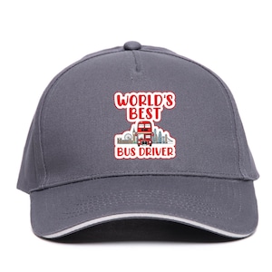 May include: A grey baseball cap with a white trim. The cap displays a red and white graphic that reads "WORLD'S BEST BUS DRIVER" with a red double-decker bus and London skyline illustration.