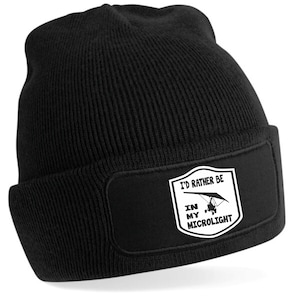 May include: Black knit beanie with a rectangular patch. The patch features the text "I'D RATHER BE IN MY MICROLIGHT" with an illustration of a microlight aircraft. The beanie is a classic, warm accessory.