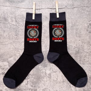 May include: Two black socks with dark grey heels and toes, suspended from a clothesline. Each sock displays a dartboard design with the text "MY LUCKY DARTS SOCKS" in red and white, encircled by a laurel wreath.