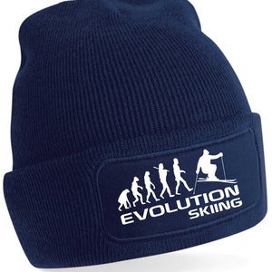 Könnte beinhalten: Eine marineblaue Strickmütze mit einem weißen Patch, der eine Silhouette der Evolution des Menschen zeigt, die in einem Skifahrer gipfelt. Der Patch lautet "Evolution Skiing".