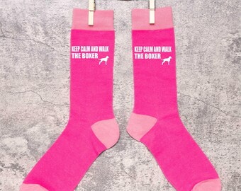 Puede incluir: Un par de calcetines rosa brillante con puños, talones y punteras de color rosa claro, colgados de pinzas de madera. El texto blanco "KEEP CALM AND WALK THE BOXER" y una silueta blanca de un perro están impresos.