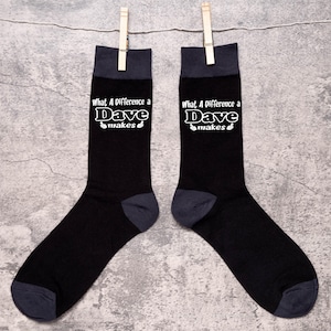 Peut inclure: Une paire de chaussettes noires avec un motif blanc qui dit "What A Difference a Dave Makes" avec des pouces en l'air de chaque côté du texte.