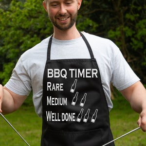 Puede incluir: Delantal de barbacoa negro con texto blanco que dice "BBQ Timer" y una lista de niveles de cocción para la carne, con botellas de cerveza como indicador de tiempo. El delantal tiene una correa blanca y lo lleva puesto una persona que está asando pollo en una barbacoa negra.