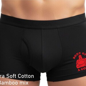 May include: Black boxer briefs featuring a wide elastic waistband with the embossed text ALPHA. The right leg displays a red graphic of a smiling figure with the text BODY OF A GOD! The fabric is labelled as an Ultra Soft Cotton Bamboo mix.