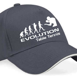 Può includere: Un berretto da baseball blu navy con una grafica bianca dell'evoluzione del tennis da tavolo. La grafica mostra una serie di figure stilizzate che evolvono da un uomo primitivo a una persona che gioca a tennis da tavolo. Il testo "EVOLUTION Tennis da tavolo" è sotto la grafica.
