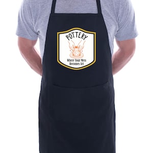Może przedstawiać: Czarny fartuch z dużą przednią kieszenią oraz paskami na szyję i talię. Fartuch posiada biało-złoty emblemat z napisem "POTTERY" i frazą "Where Your Mess Becomes Art" czarnym tekstem, a także ilustracją dłoni formujących glinę.