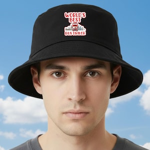 May include: Black bucket hat featuring a red and white graphic that reads "WORLD'S BEST BUS DRIVER". The graphic includes a red bus and a city skyline. The hat is worn by a person.