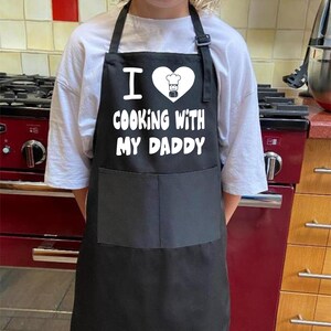 May include: Black apron with a white heart and the text "I love cooking with my daddy".