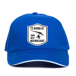 May include: A royal blue baseball cap with a white trim and a patch that reads "I'D RATHER BE IN MY MICROLIGHT." The patch features a black and white illustration of a microlight aircraft. The cap is made of a durable fabric.