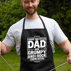 Puede incluir: Delantal negro con texto blanco: "I HAVE TWO TITLES DAD AND GRUMPY AND I ROCK THEM BOTH". El delantal se usa mientras se asa comida a la parrilla.