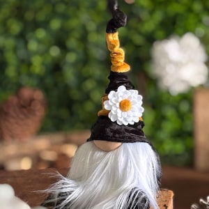 May include: A black and white gnome with a long white beard. The gnome is wearing a black velvet hat with a yellow velvet band and a white felt flower.