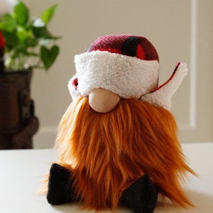 May include: A plush gnome with a red and black checked hat, a white fluffy beard, and a white fluffy eye mask. The gnome is sitting on a white surface.