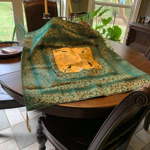 May include: A green and gold patterned tablecloth with a gold tassel, draped over a dark wooden table. The tablecloth has a central gold square with a floral design. The table is surrounded by dark wooden chairs.