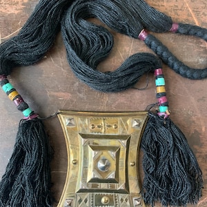 May include: A gold-colored metal pendant with intricate geometric designs and a central silver-colored circle. The pendant is attached to a long black cord with tassels on each end. The cord is decorated with colorful beads.