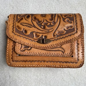 May include: A tan leather purse with intricate floral tooling and a braided leather trim. The purse has a brass clasp and a flap closure.