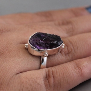Op de afbeelding: Een zilveren ring met een grote, ruwe amethist edelsteen. De amethist is diep paars van kleur en heeft een ruwe, natuurlijke textuur.