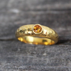Könnte beinhalten: Ein goldener Bandring mit gehämmerter Textur und einem kleinen, runden, orangefarbenen Edelstein, der in der Mitte eingesetzt ist.